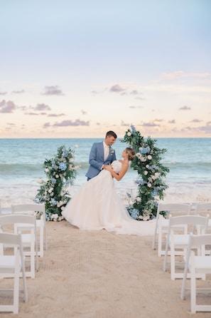 A beach wedding ceremony with a floral arch and white chairs.