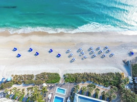 An aerial view of a beach with rows of blue umbrellas and lounge chairs, a pool area with a clear blue pool, and a building with a sloped roof.