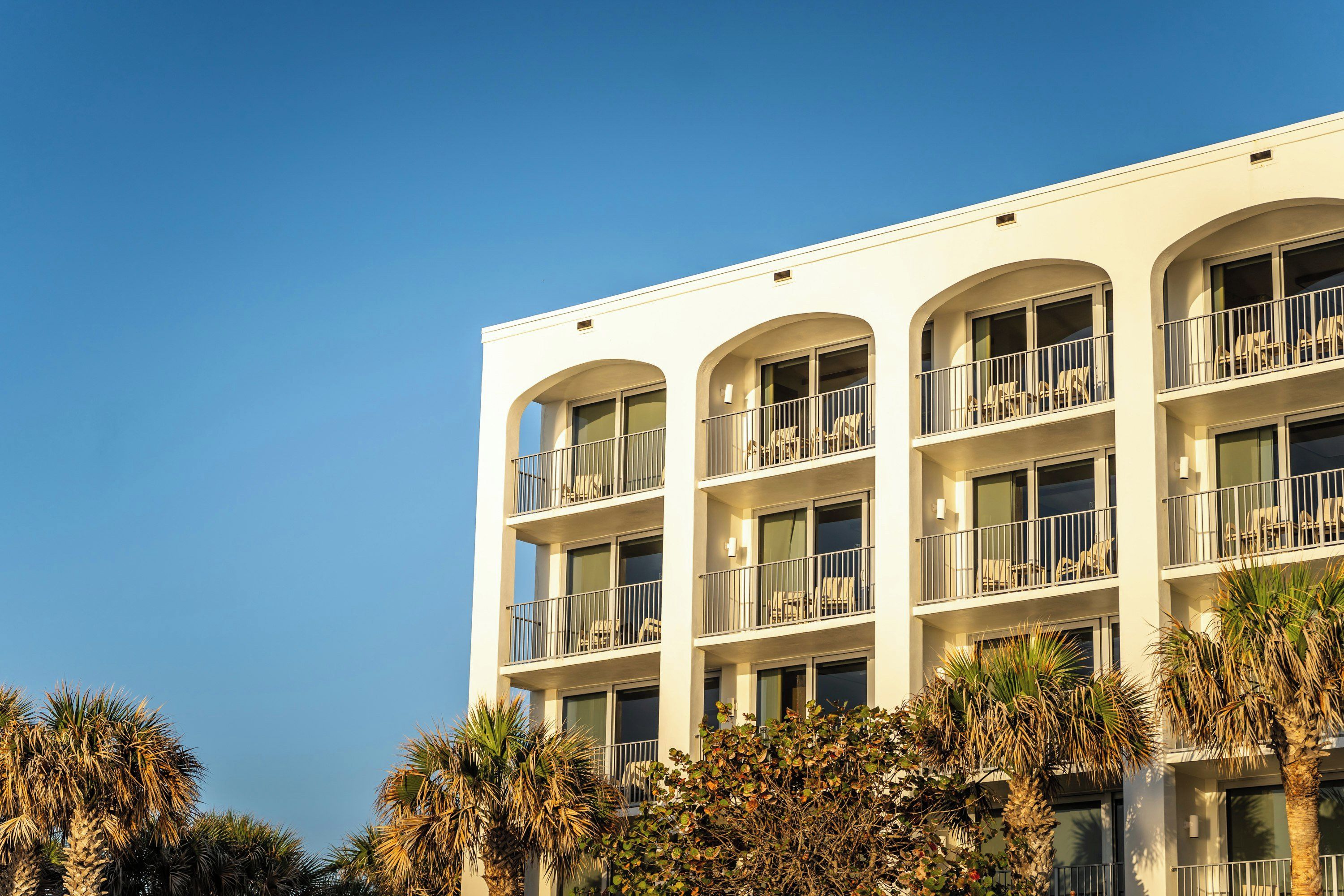 A multi-story hotel with balconies, large windows, and palm trees in the foreground.