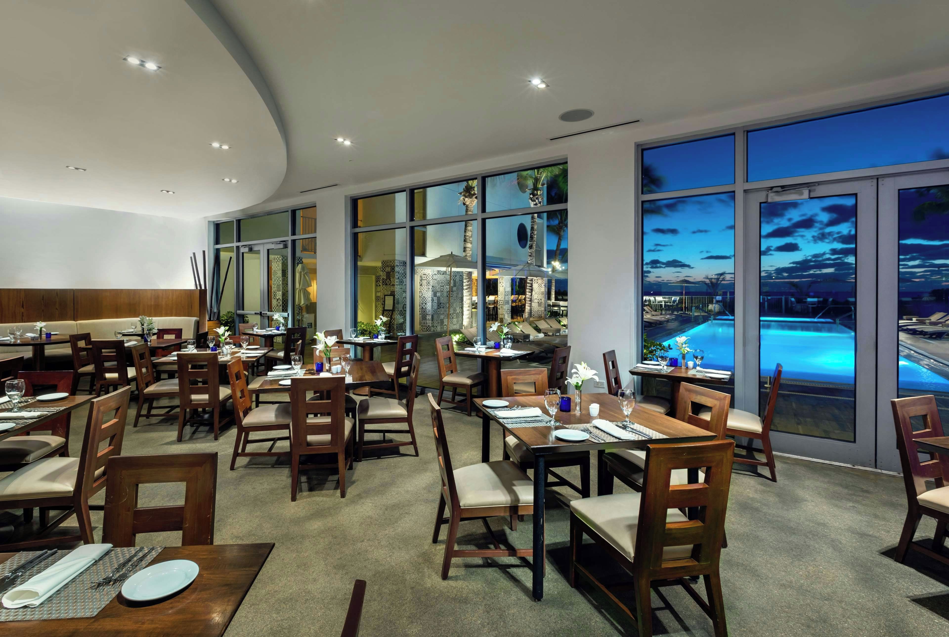 A restaurant with multiple dining tables and chairs, large windows with a view of a pool and sunset, and a modern interior design.