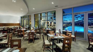 Ein Restaurant mit mehreren Tischen und Stühlen, großen Fenstern mit Blick auf einen Pool und Sonnenuntergang sowie einem modernen Innenraum.