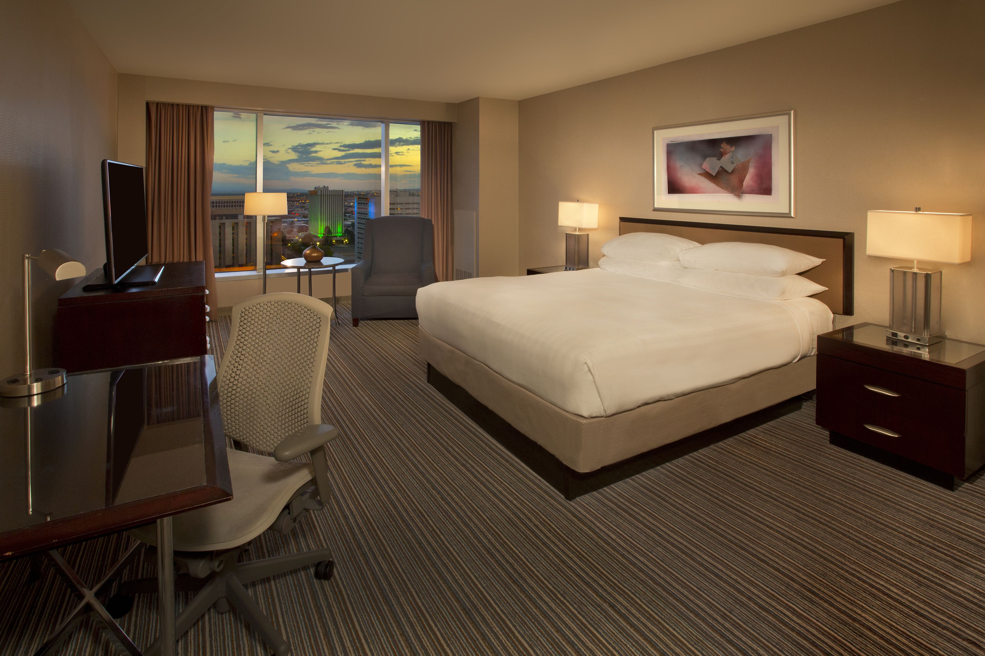 A hotel room with a large bed, a desk with a computer, a chair, two bedside tables with lamps, and a window with a view of the city.