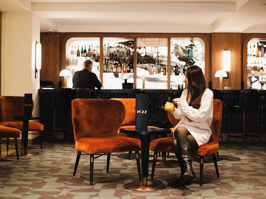 Uma área de bar com uma mulher sentada a uma mesa, um bartender ao fundo e um balcão de bar com vários frascos.