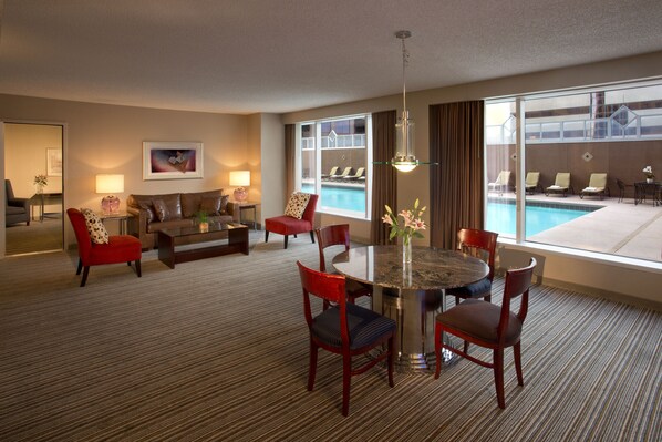 A hotel room with a dining area, a sofa, a red armchair, a coffee table, and a view of a swimming pool.