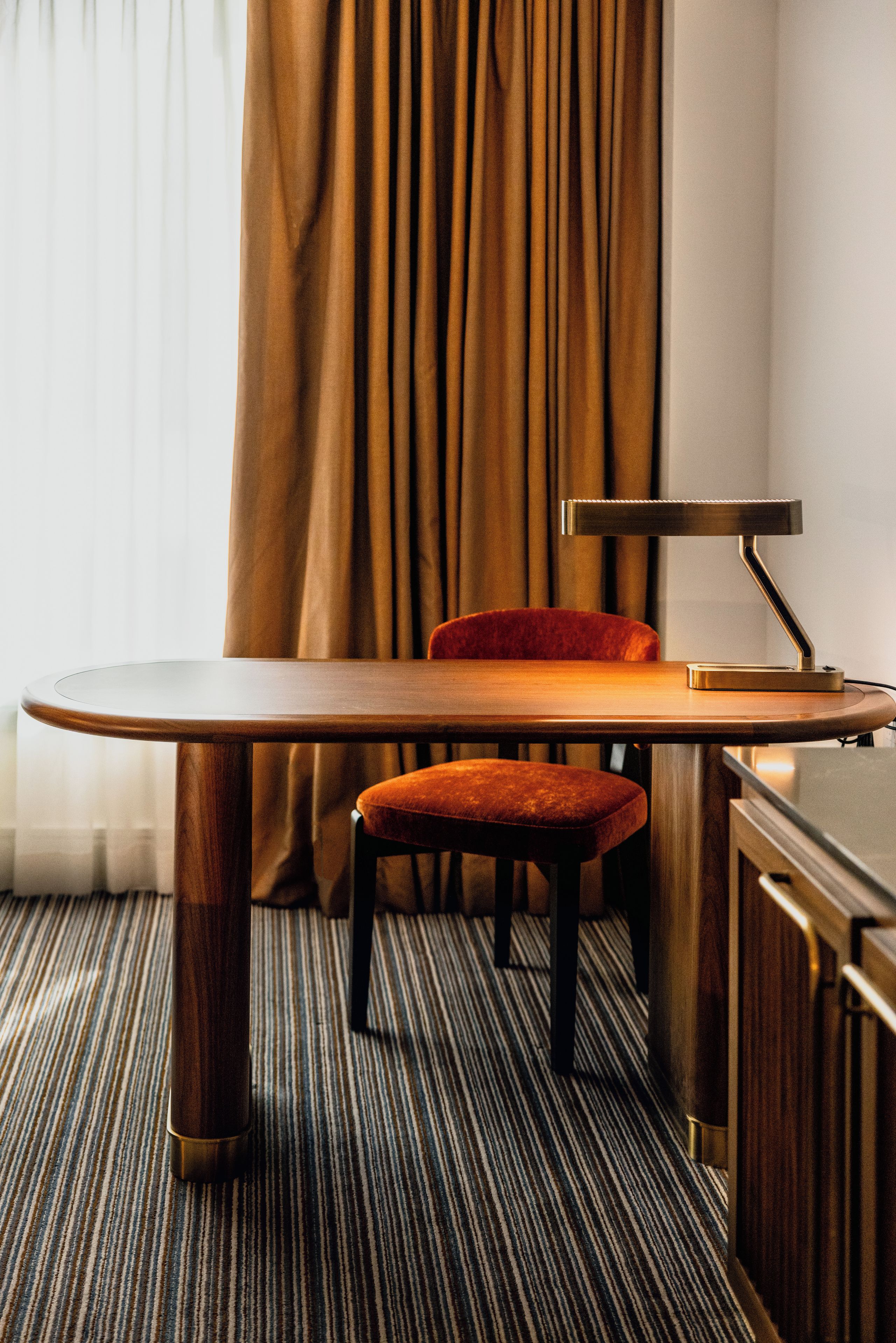 A hotel room with a wooden table, a red upholstered chair, a desk lamp, and a striped carpet.