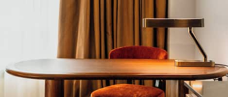 A hotel room with a wooden table, a red upholstered chair, a desk lamp, and a striped carpet.