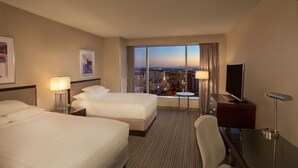 Habitación de hotel con dos camas, un escritorio, una silla, un televisor y un ventanal con vistas a la ciudad.