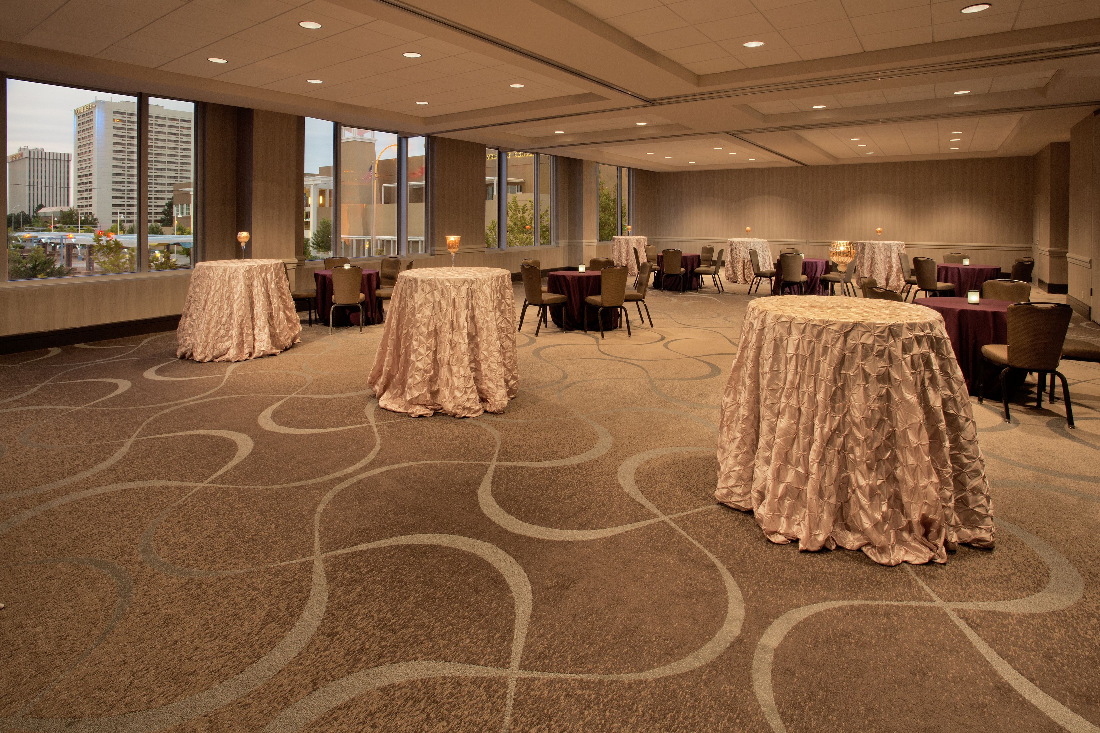 A spacious conference room with large tables covered in tablecloths, arranged in rows, and large windows offering a view of the cityscape.