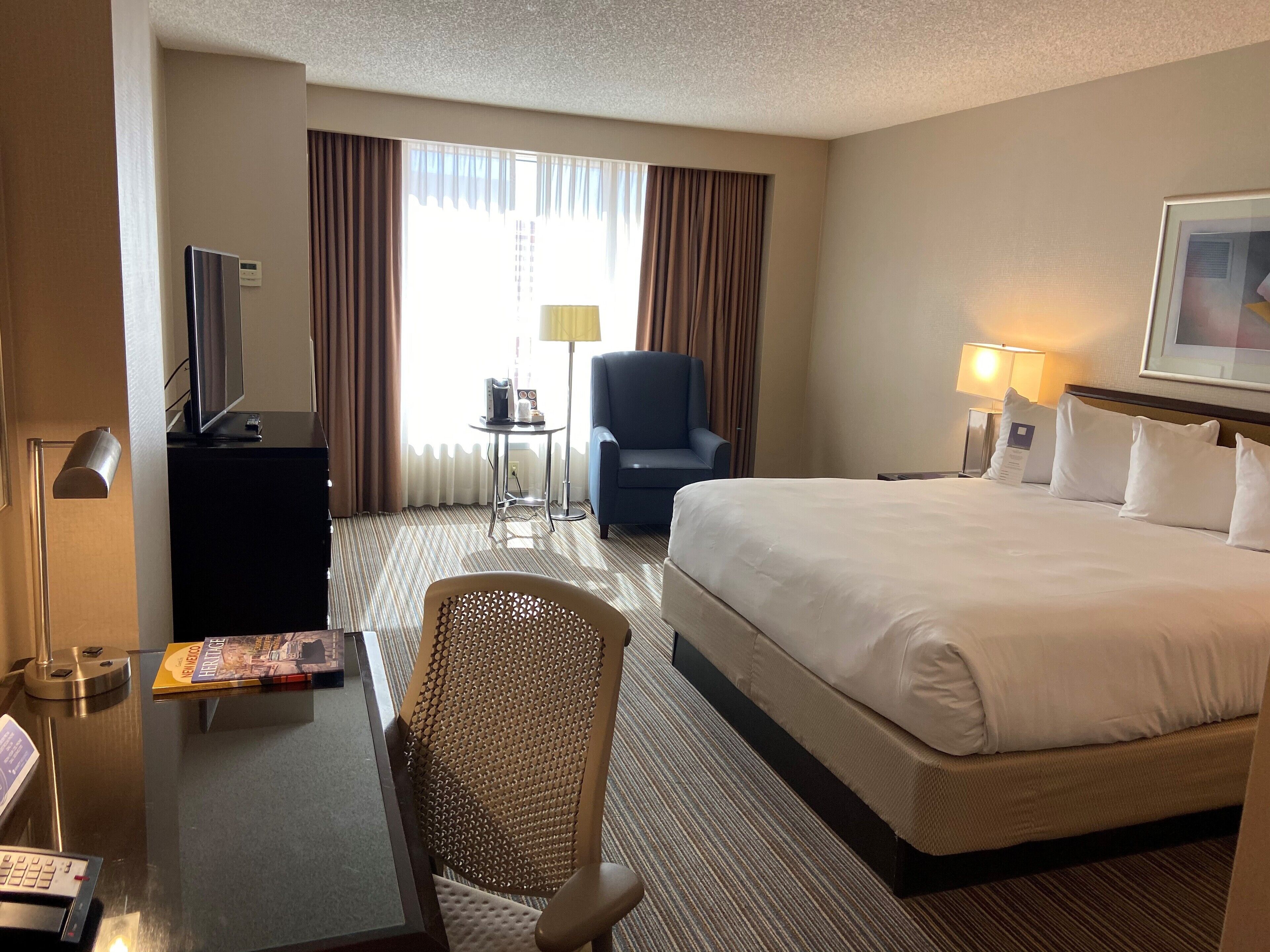 A hotel room with a large bed, a desk with a computer, a chair, a small table with a lamp, and a window with curtains.