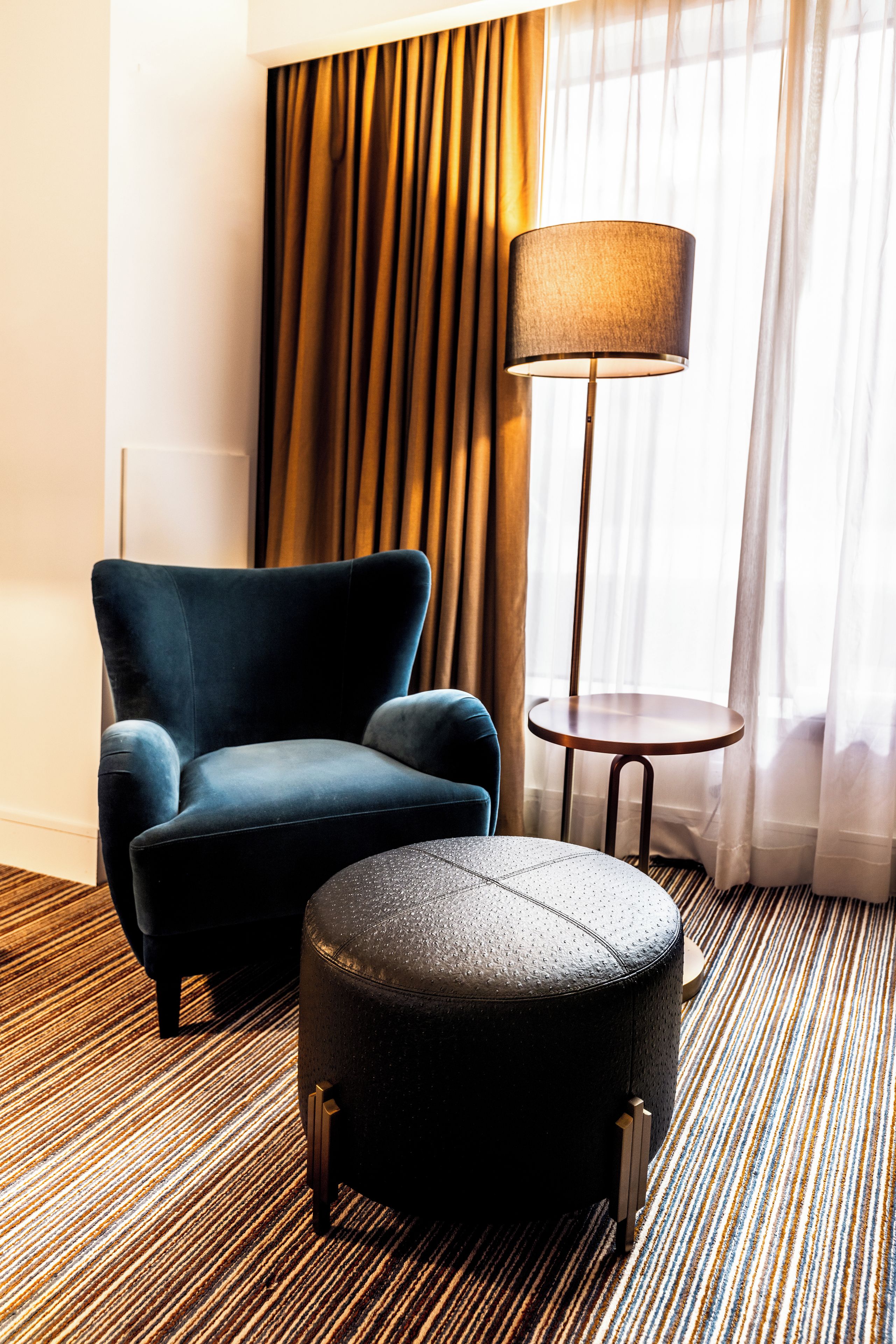 A hotel room with a dark blue armchair, a round ottoman, a floor lamp, and a striped carpet.