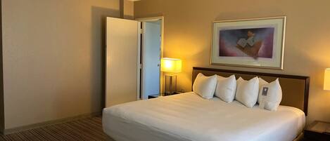 A hotel room with a large bed, a door, a framed picture on the wall, and a nightstand with a lamp.