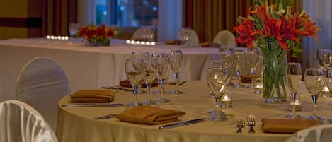 A banquet table set for a formal event with a white tablecloth, multiple wine glasses, and a centerpiece of red flowers.
