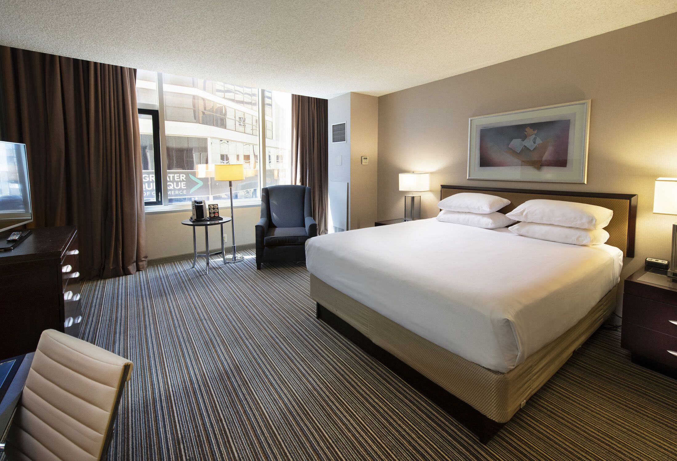 A hotel room with a large bed, a desk with a chair, a small table, and a window with curtains.