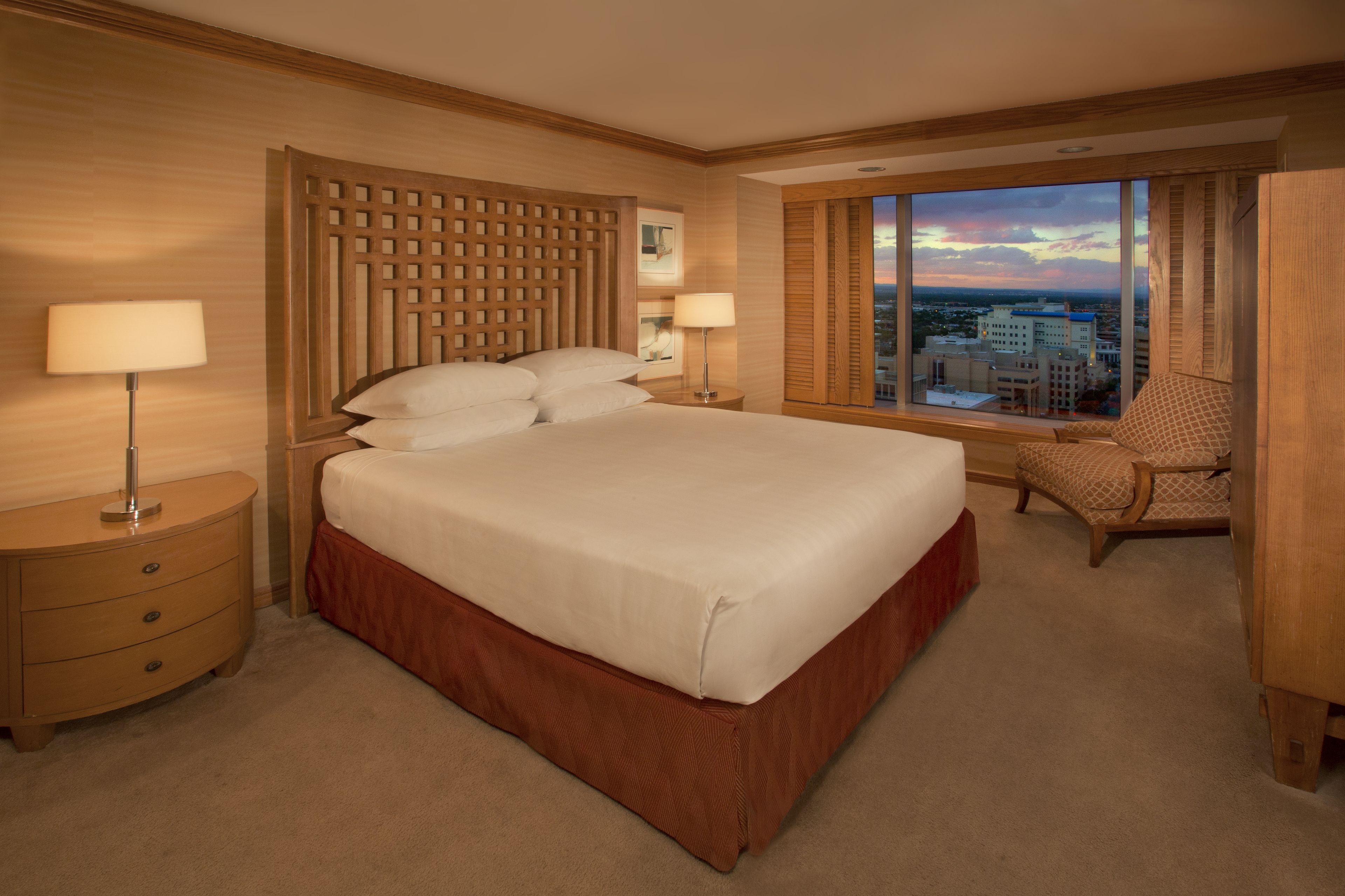 A hotel room with a large bed, two bedside tables with lamps, a wooden headboard, a city view through large windows, and a chair.