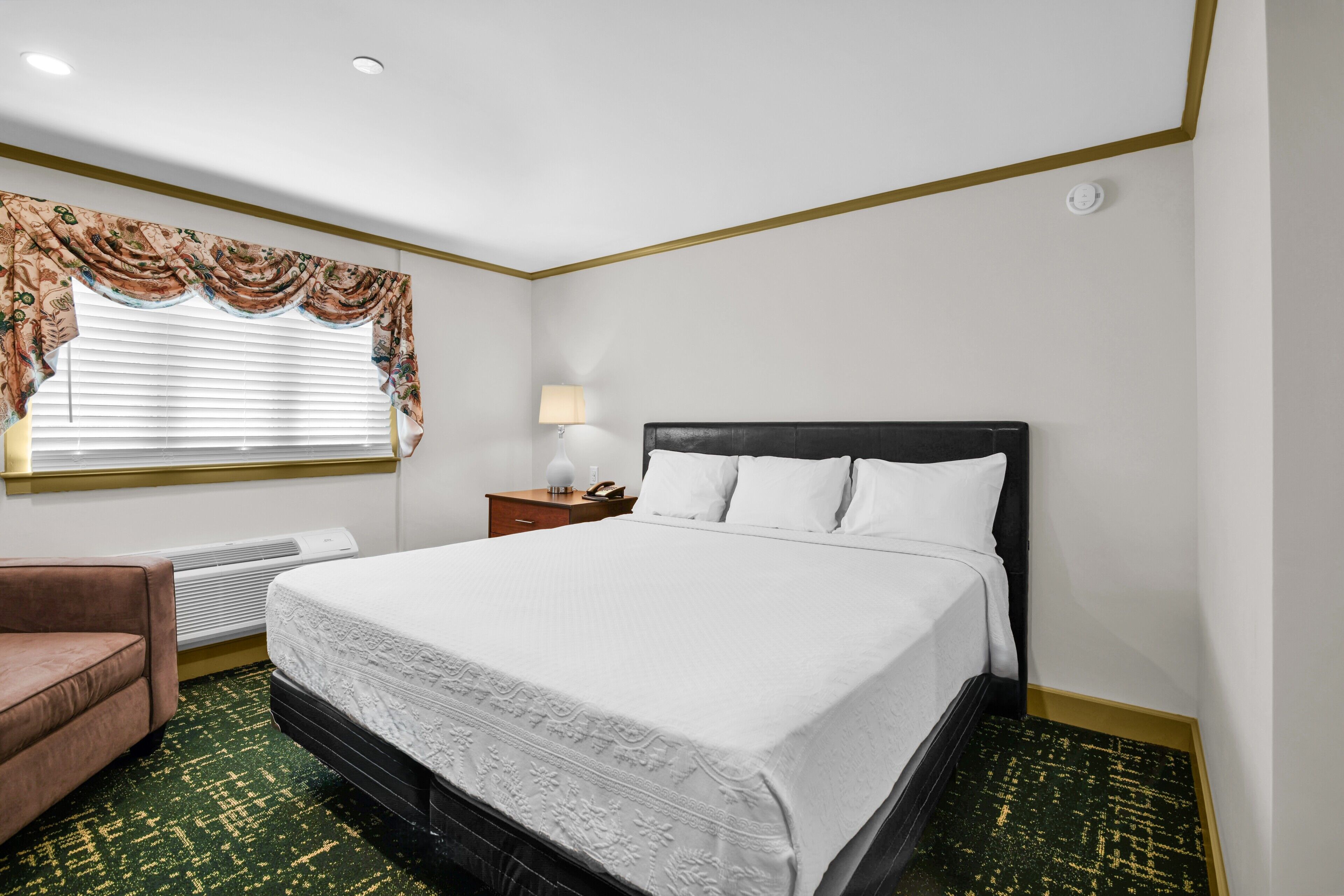 Quarto de hotel com cama, poltrona, mesa de cabeceira, luminária e janela com persianas.