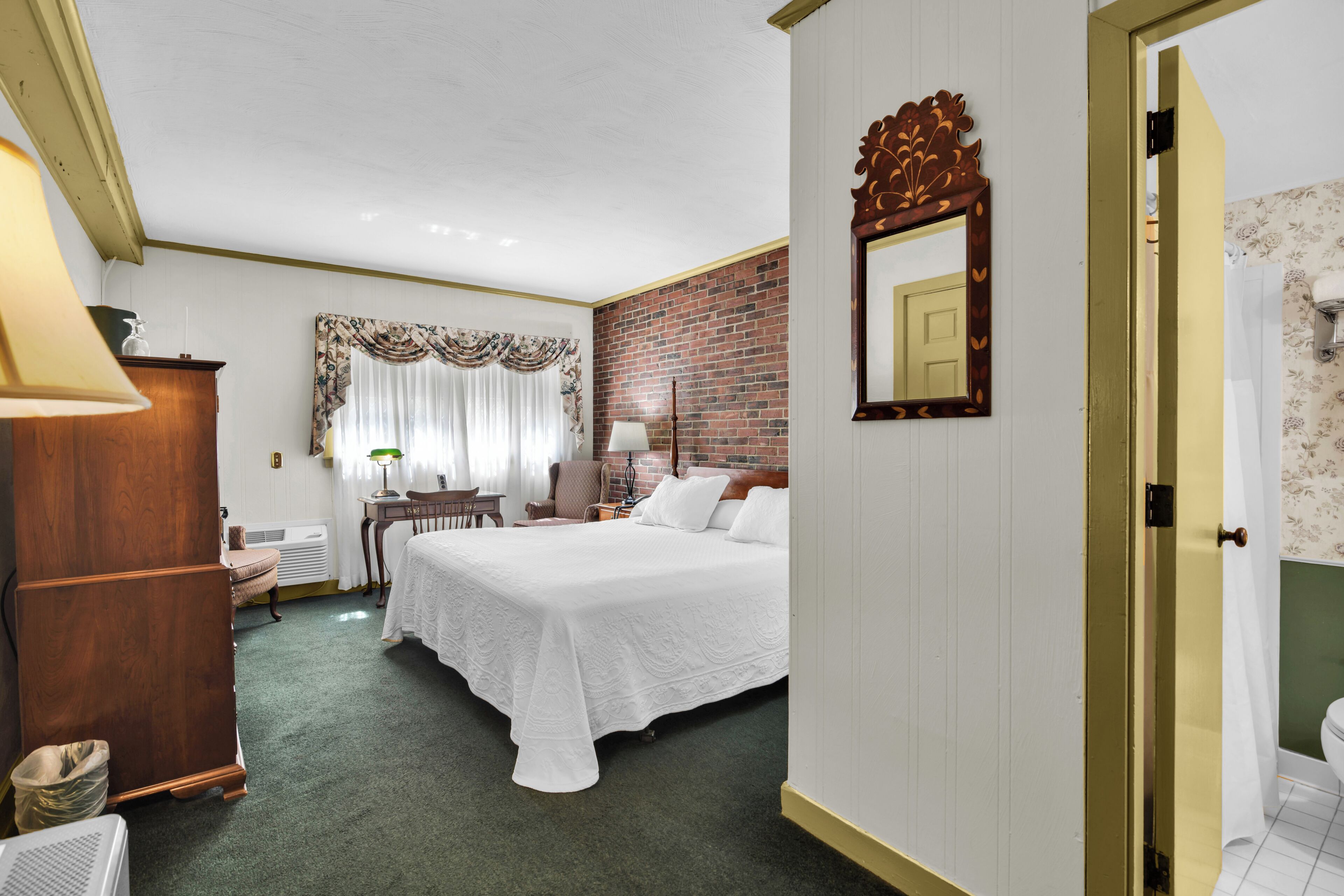 Habitación de hotel con cama, escritorio, silla, lámpara, espejo y puerta que da a un baño.