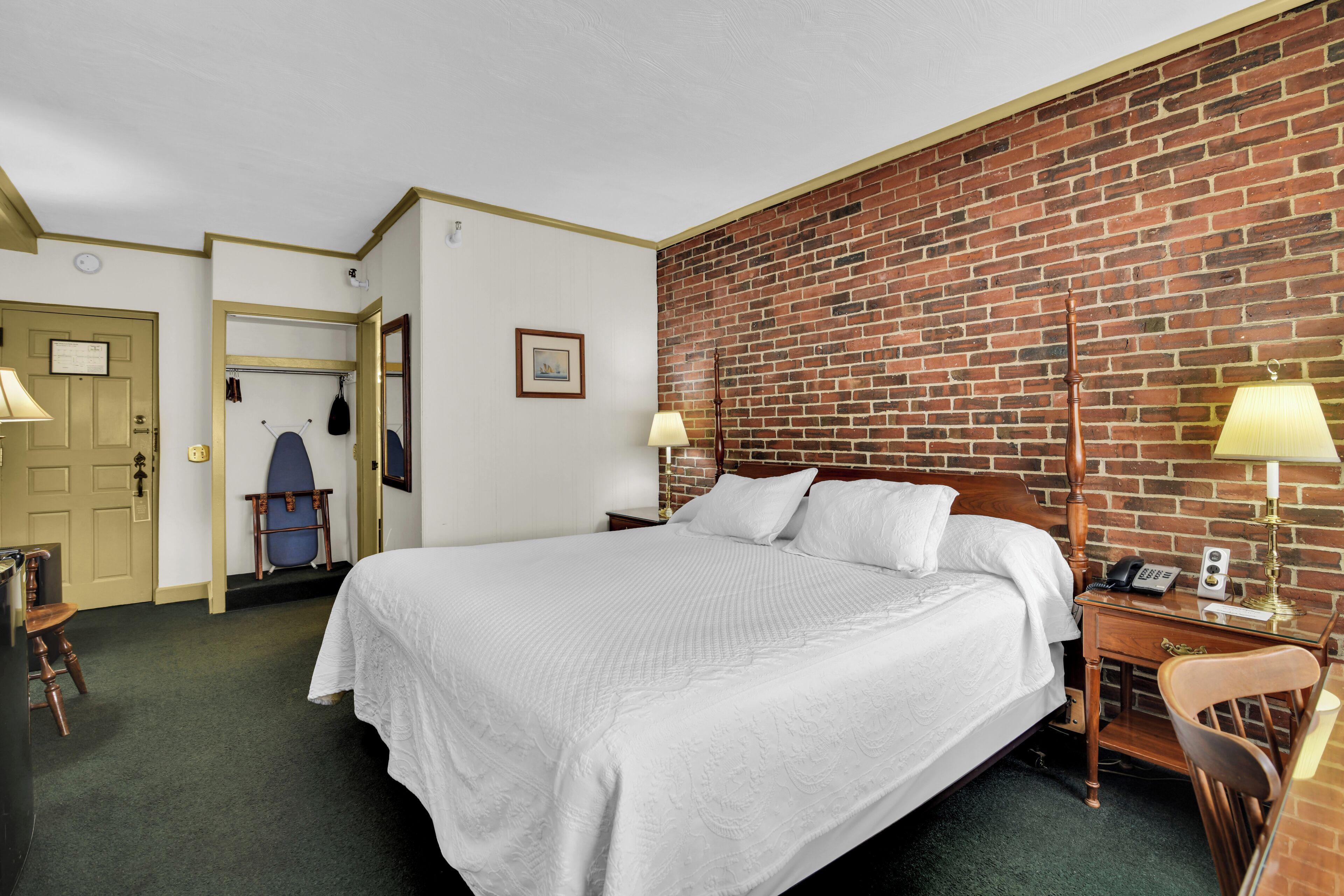 A hotel room with a large bed, a desk with a lamp, a chair, a coat rack, and a brick wall.