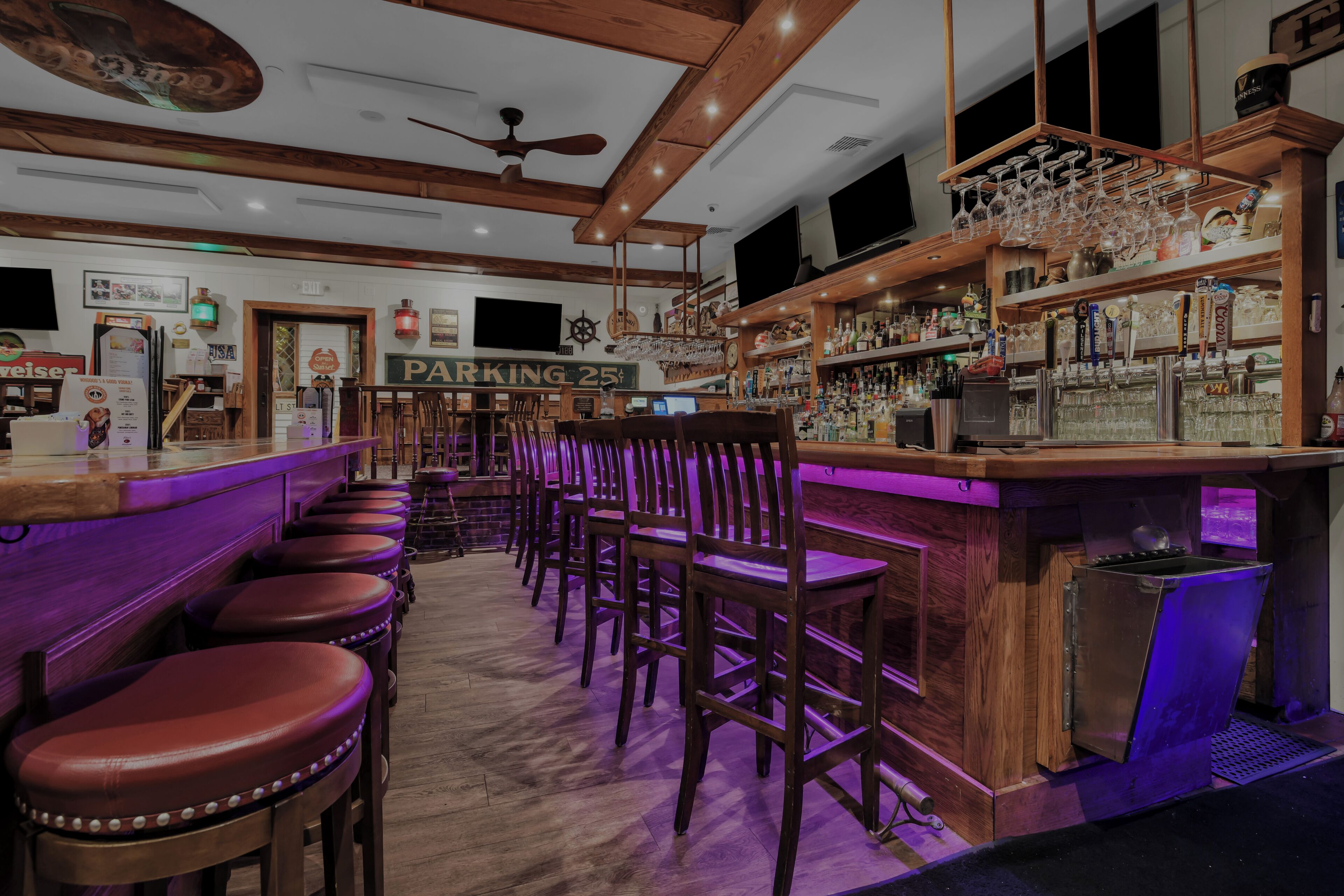 Una zona de bar con taburetes de madera, un mostrador con un letrero de neón y una variedad de botellas y vasos.