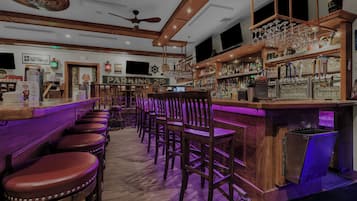 Una zona de bar con taburetes de madera, un mostrador con un letrero de neón y una variedad de botellas y vasos.