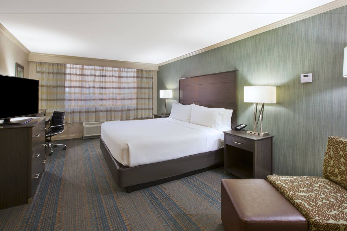 A hotel room with a large bed, a desk with a TV, two bedside lamps, a chair, and a patterned sofa.