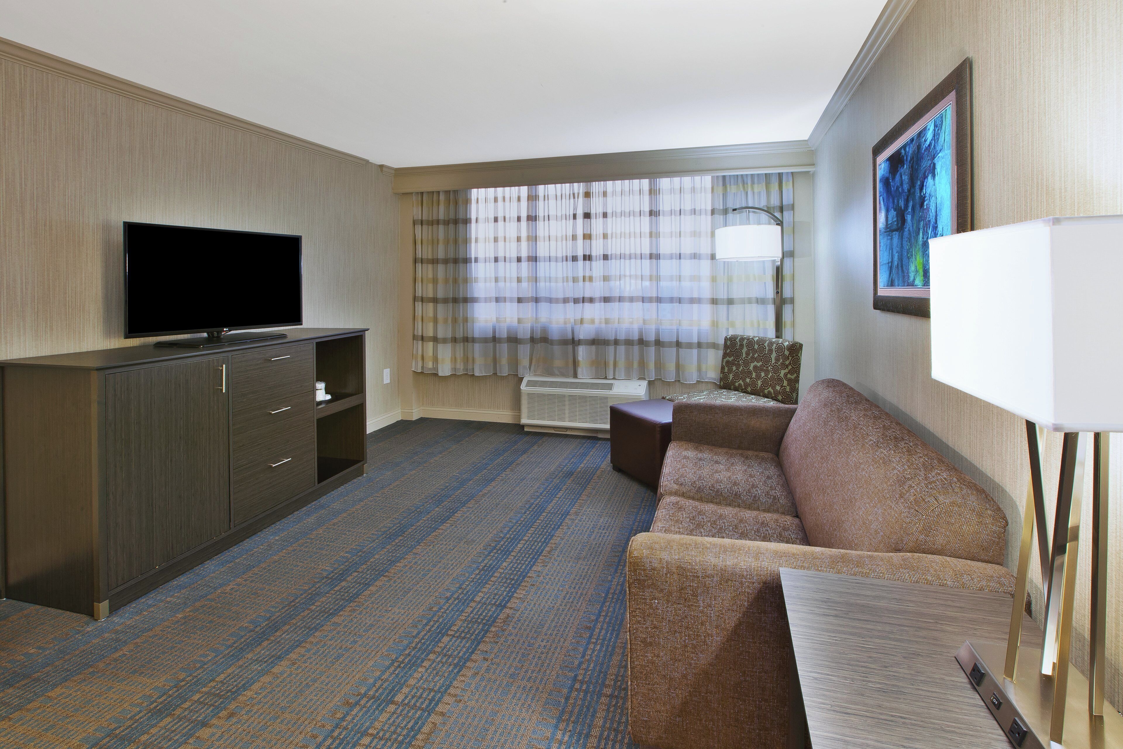 A hotel room with a flat-screen TV, a sofa, a small table, and a window with curtains.