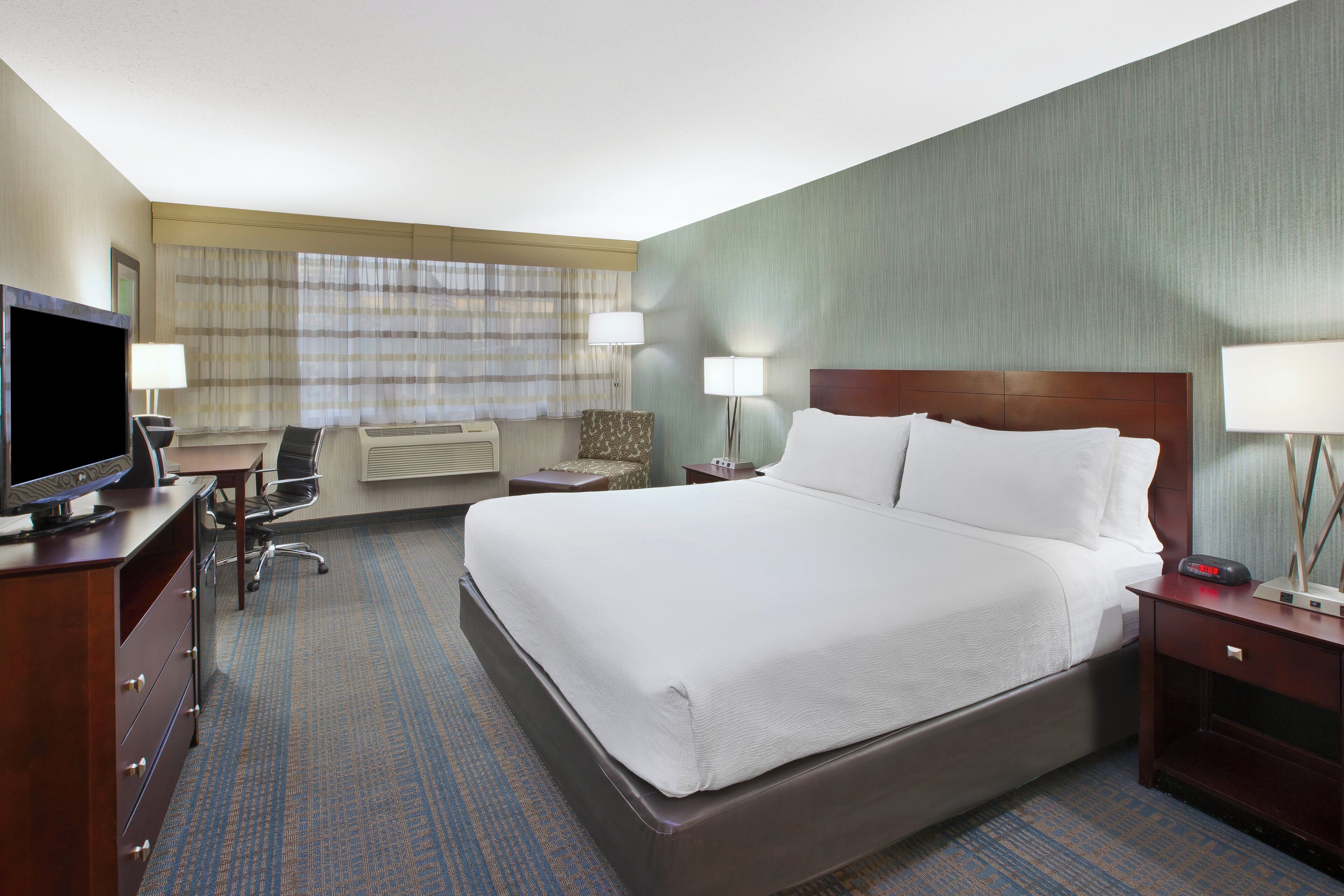 A hotel room with a large bed, a desk with a television, two bedside lamps, a chair, and a small table.