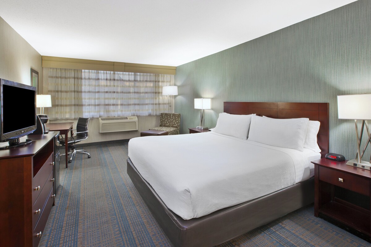 A hotel room with a large bed, a desk with a television, two bedside lamps, a chair, and a small table.