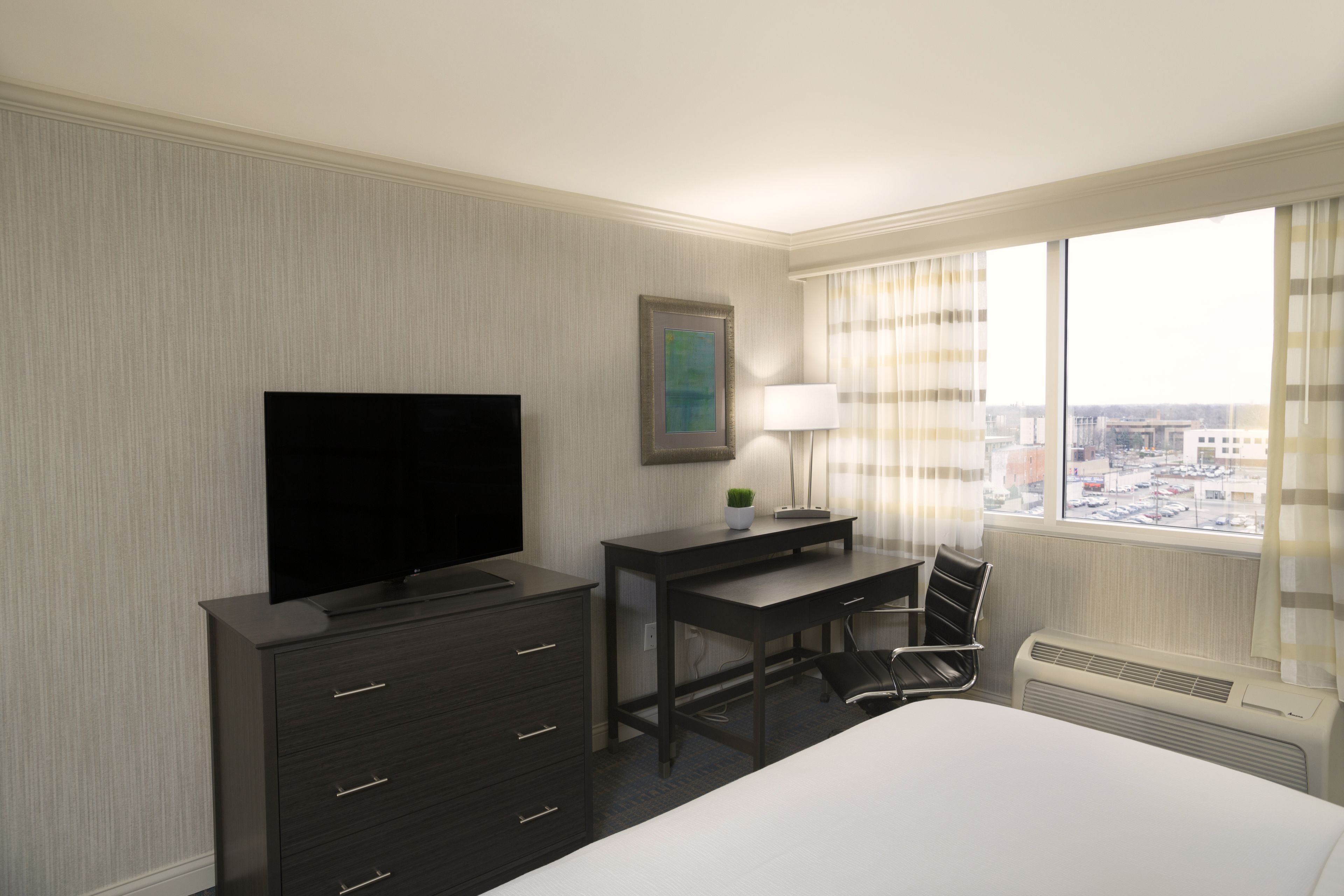 A hotel room with a bed, a desk with a chair, a television, a desk lamp, a potted plant, and a window with curtains.