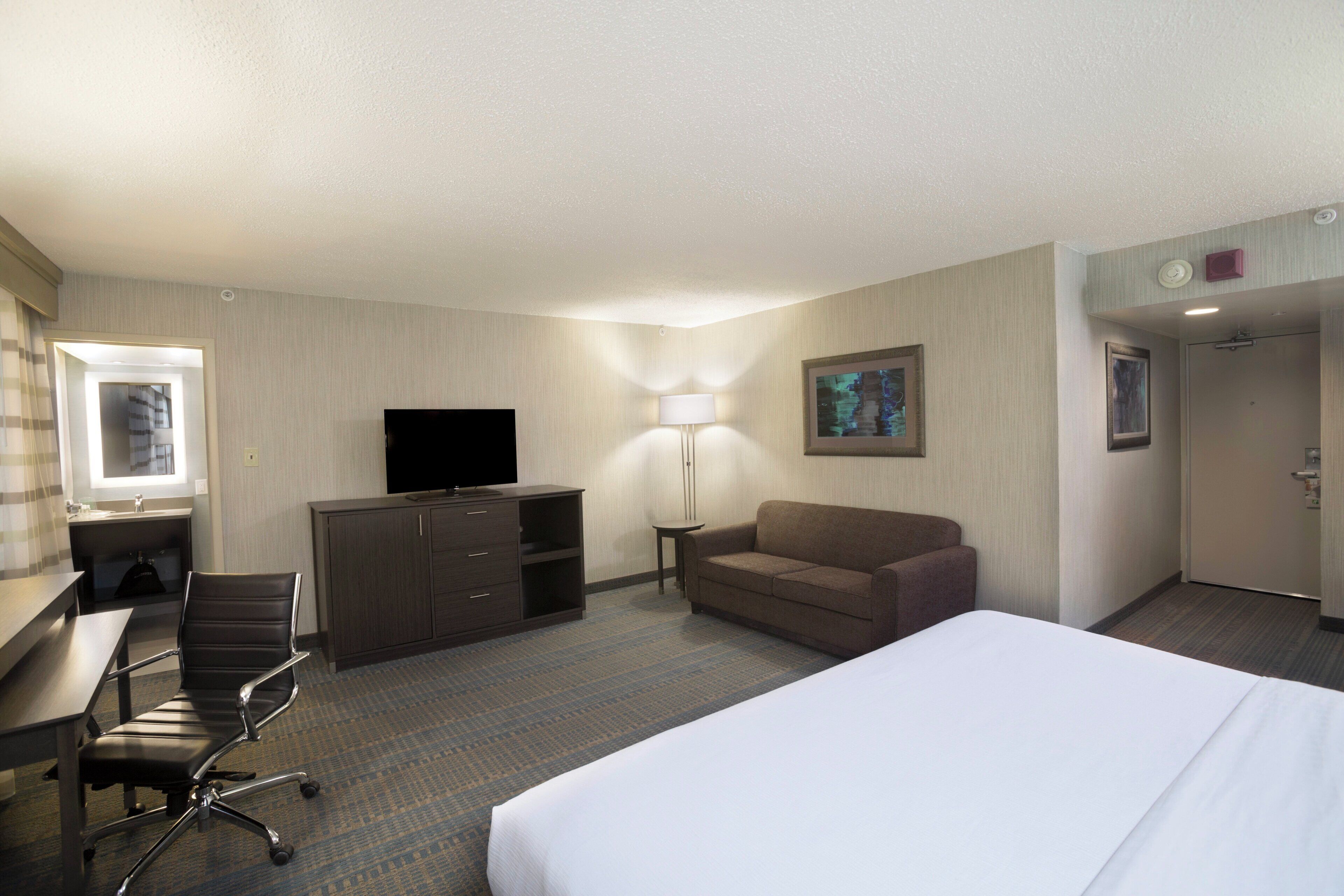 A hotel room with a bed, a desk with a chair, a television, a sofa, and a lamp.