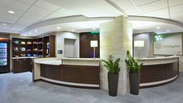 A hotel reception area with a curved counter, a refrigerator stocked with snacks, and two potted plants.