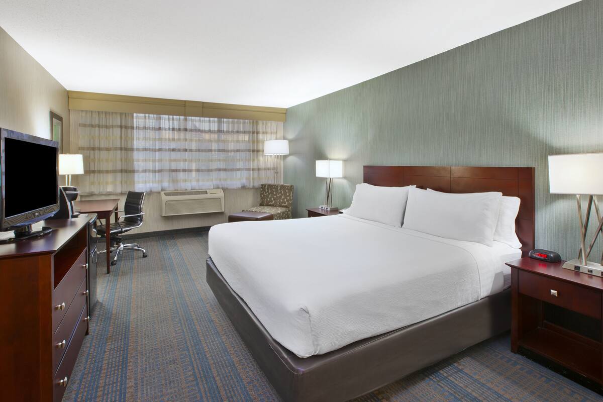 A hotel room with a large bed, a desk with a flat-screen TV, two bedside tables with lamps, a chair, and a carpeted floor.