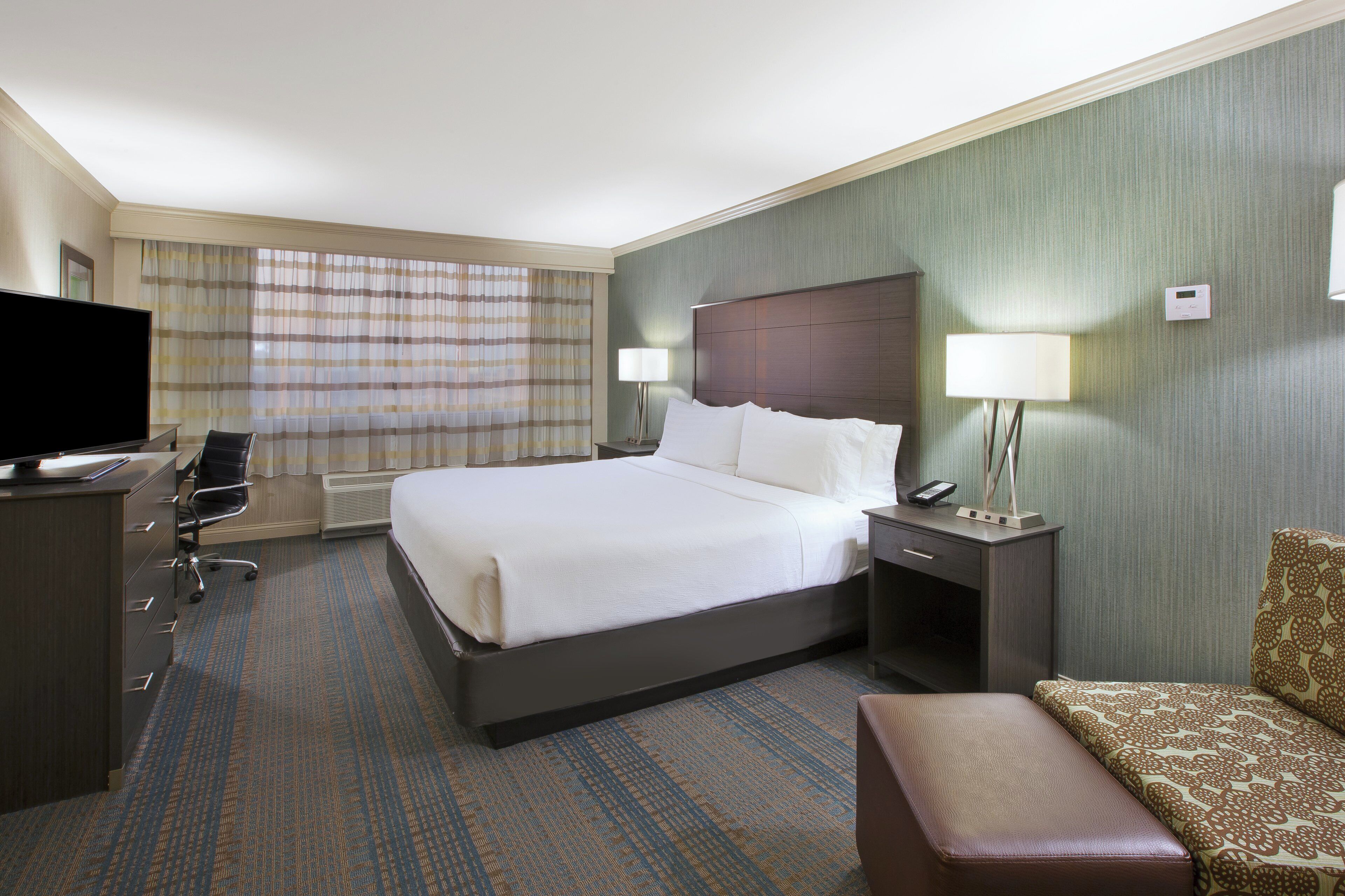 A hotel room with a bed, a desk with a TV, a chair, a bedside table with lamps, and a patterned sofa.