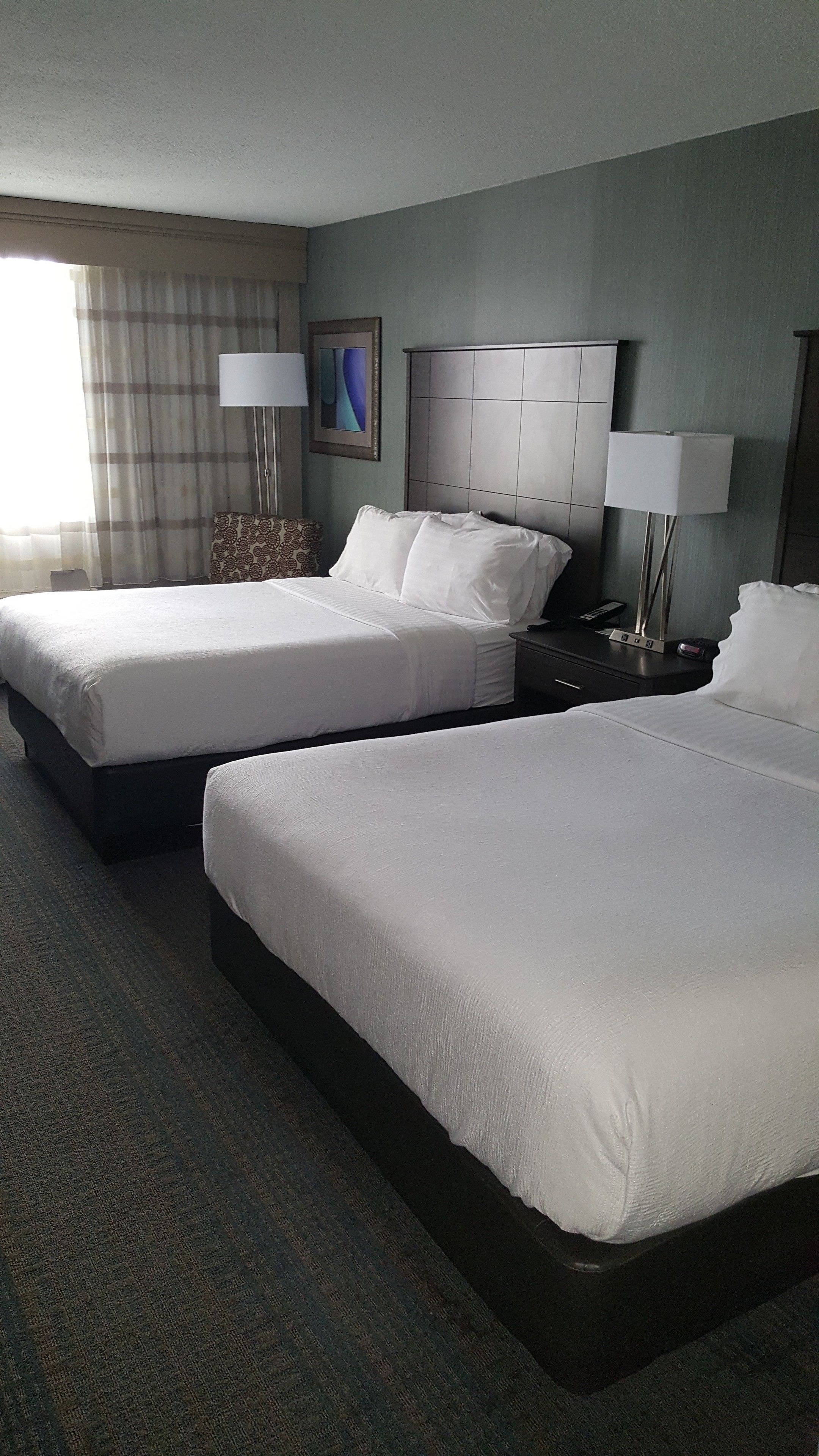 A hotel room with two beds, a headboard, bedside tables, lamps, a painting, and a window with curtains.