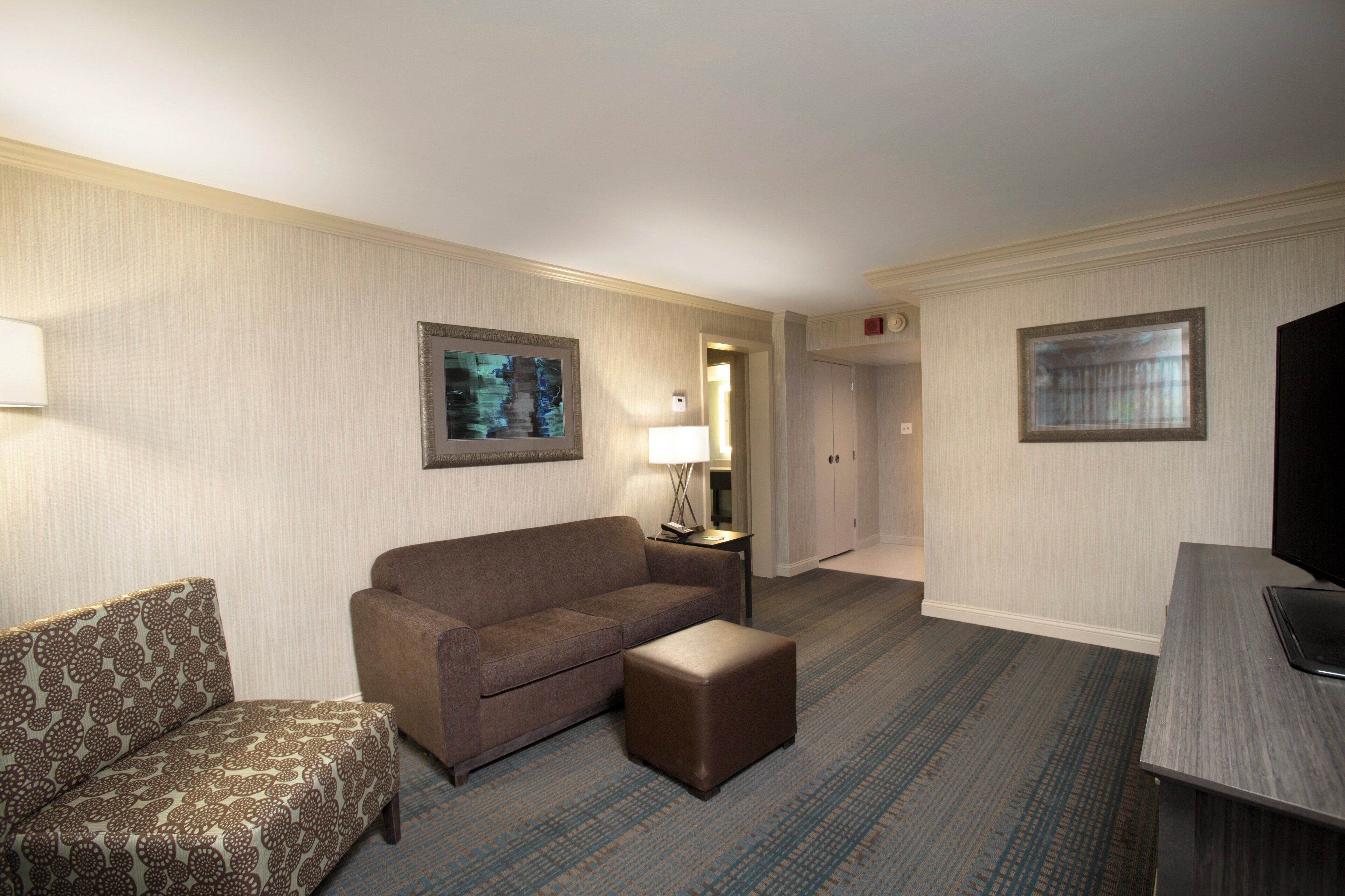 A hotel room with a sofa, ottoman, a flat-screen TV, a lamp, and two framed pictures on the walls.