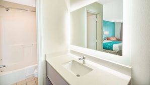 Ein Badezimmer mit Badewanne, Toilette, Waschbecken und einem Spiegel, der ein Schlafzimmer mit Bett und Nachttisch reflektiert.