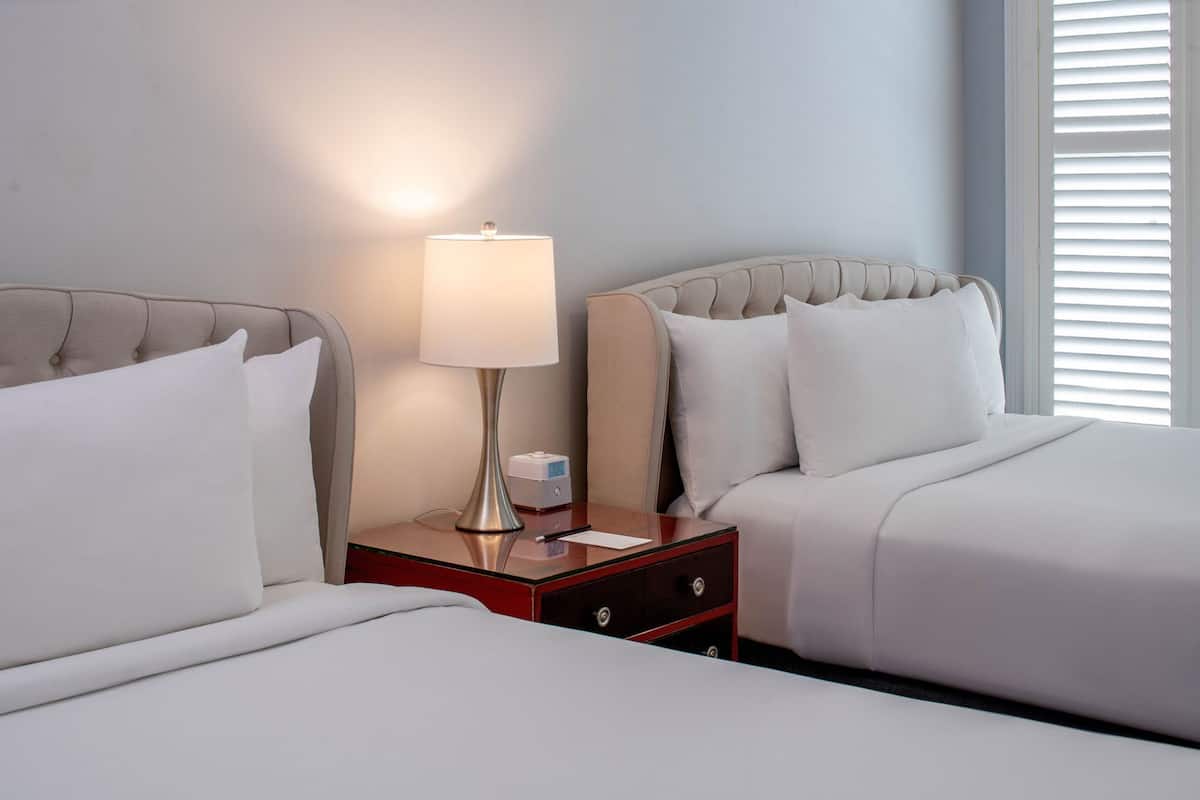 A hotel room with two beds, a bedside table with a lamp, and a window with blinds.