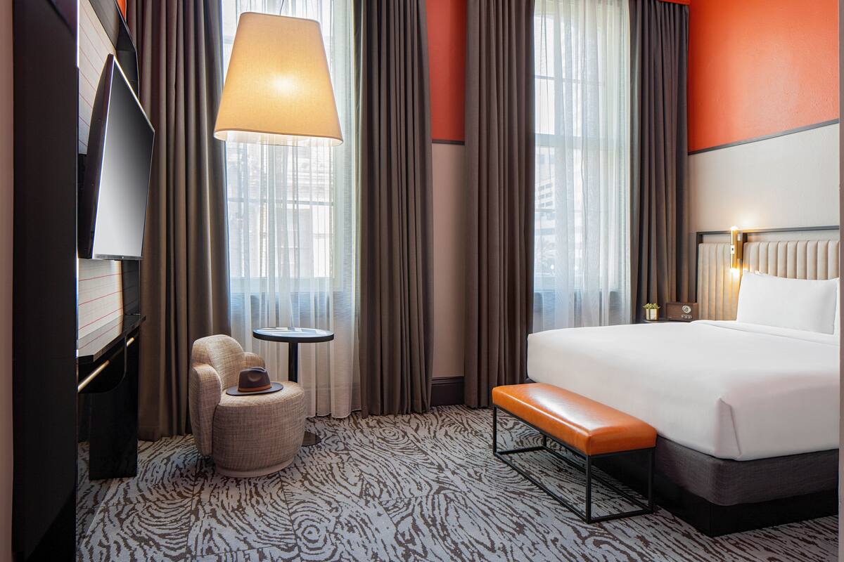 A hotel room with a bed, a desk, a chair, a lamp, and a window with curtains.