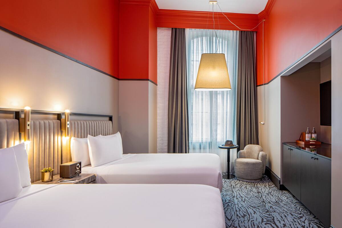 A hotel room with two beds, a desk with a lamp, a chair, a minibar, and a large window with curtains.