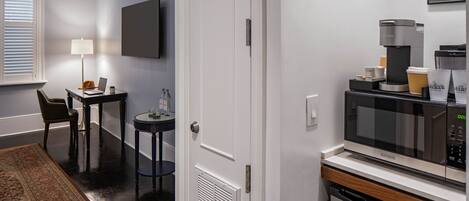 A compact hotel room with a kitchenette, a dining area with a table and chairs, a standing lamp, and a large window with blinds.