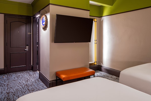 A hotel room with a large bed, an orange bench, a flat-screen TV mounted on the wall, and a door with a keypad lock.