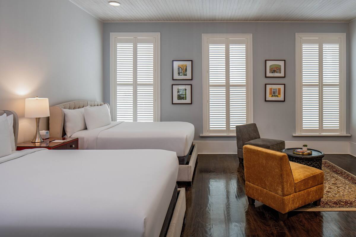 A hotel room with two beds, a chair, a table, and framed pictures on the wall.