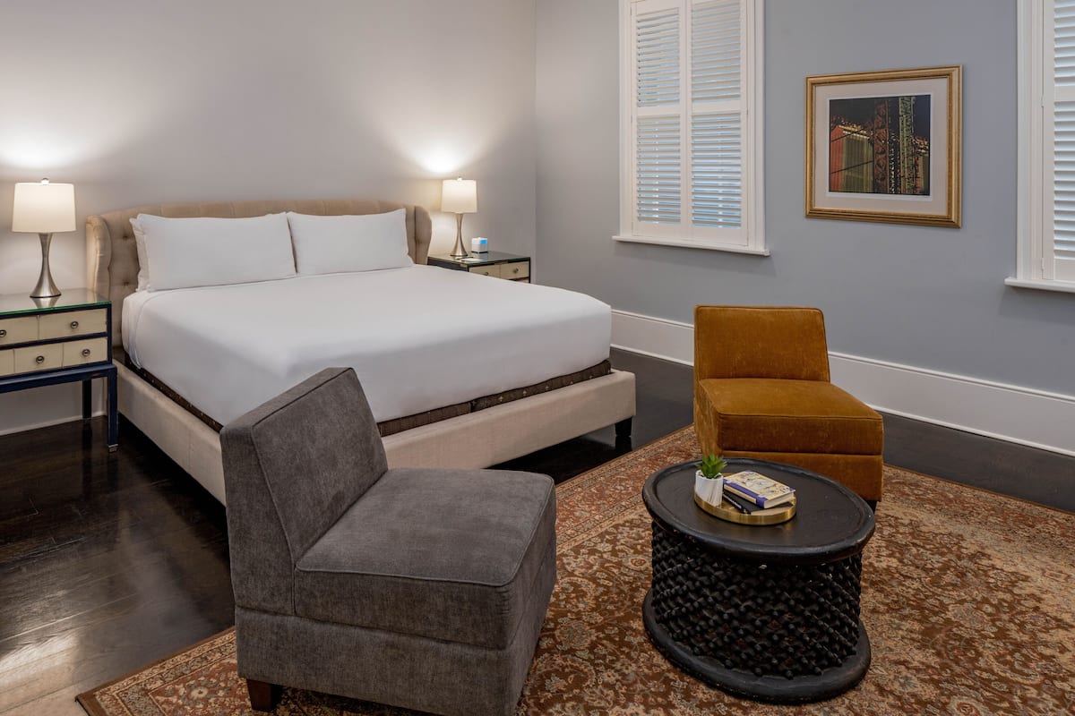 A bedroom with a bed, two bedside tables with lamps, a grey armchair, a round coffee table with a book and a plant, and a framed picture on the wall.