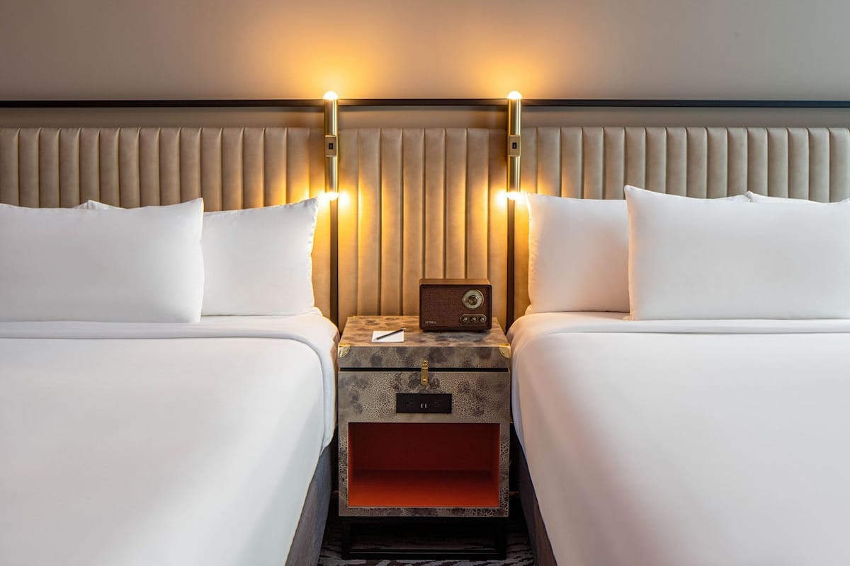 A hotel room with two beds, a headboard with vertical slats, and a nightstand with a radio and a lamp.