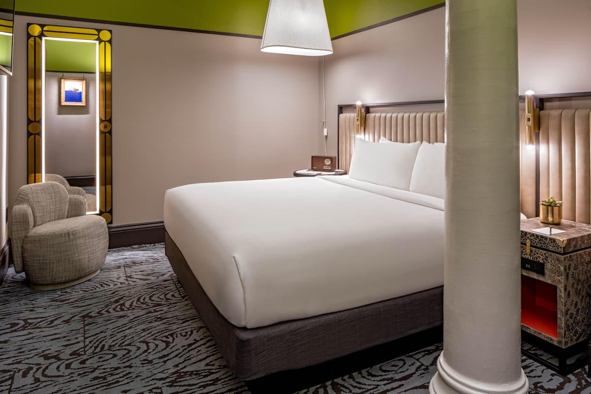 A hotel room with a large bed, a nightstand, a lamp, a chair, and a decorative mirror.