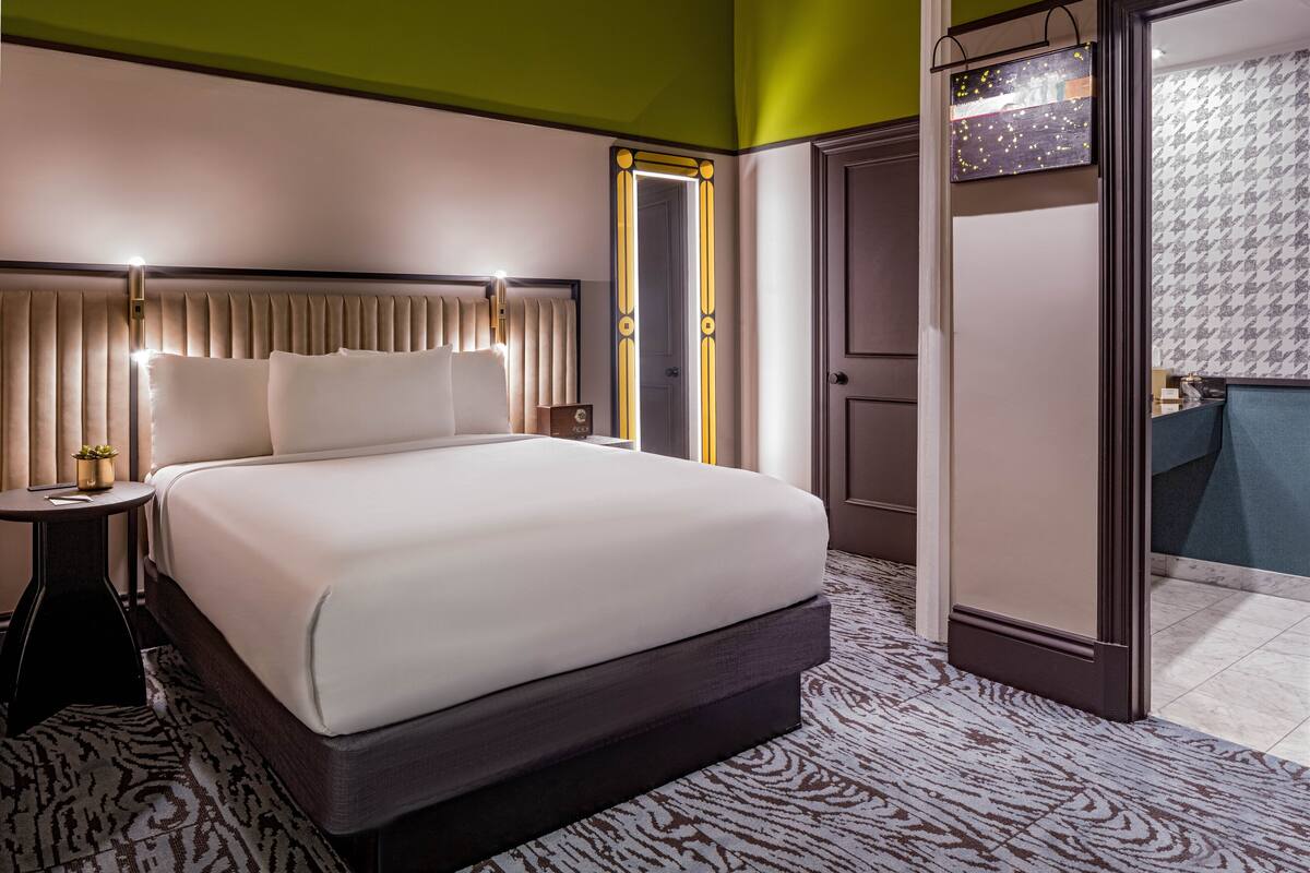 A hotel room with a large bed, bedside tables, a nightstand, a mirror, a wardrobe, and a bathroom with patterned tiles.