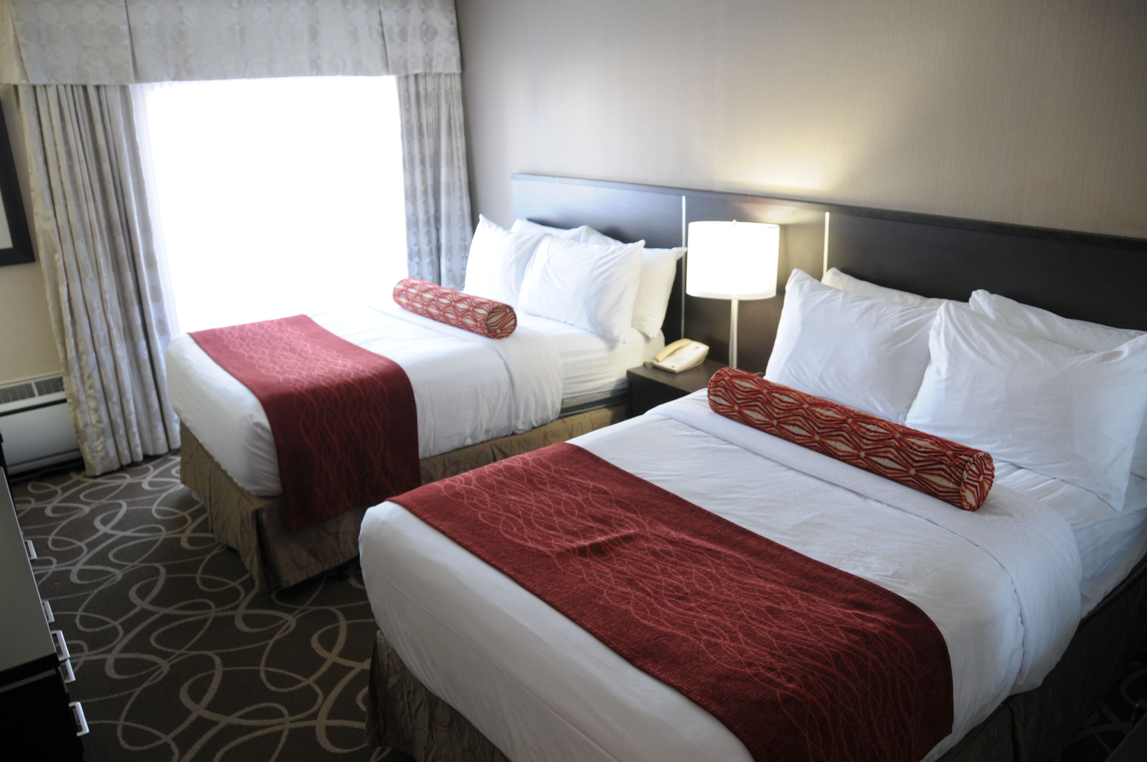 Hotelkamer met twee bedden, elk voorzien van witte lakens en rode sierkussens. Er staat een nachtkastje met een lamp en een telefoon. De kamer heeft een kamerbreed tapijt en een raam met gordijnen.