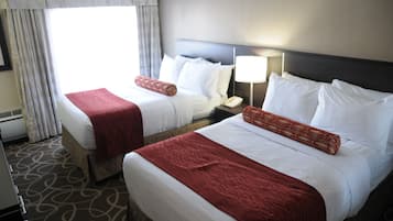 Hotelkamer met twee bedden, elk voorzien van witte lakens en rode sierkussens. Er staat een nachtkastje met een lamp en een telefoon. De kamer heeft een kamerbreed tapijt en een raam met gordijnen.