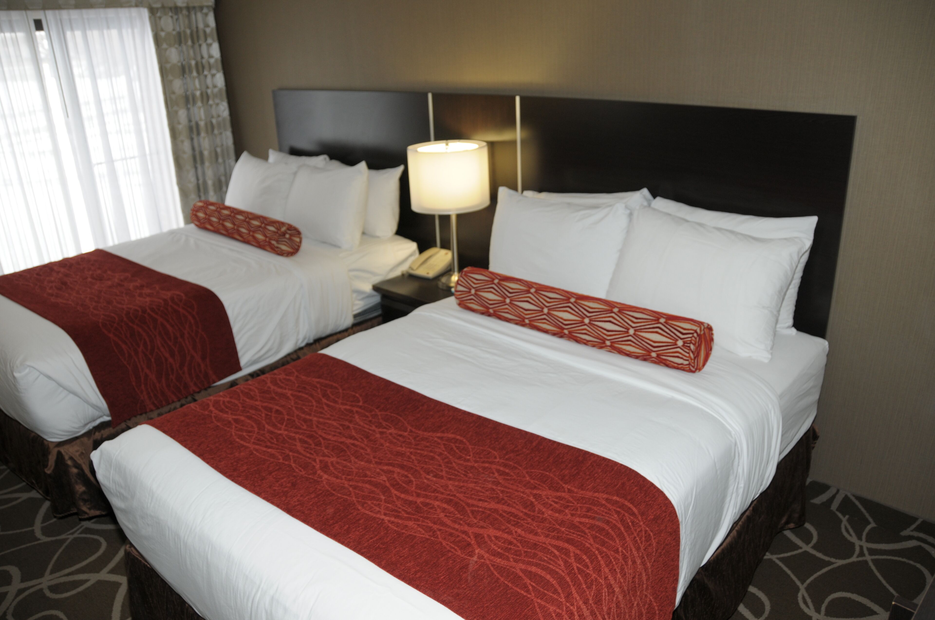 Quarto de hotel com duas camas, cada uma com lençóis brancos e almofadas decorativas vermelhas. Há uma mesa de cabeceira com uma luminária e um telefone.