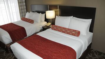 Quarto de hotel com duas camas, cada uma com lençóis brancos e almofadas decorativas vermelhas. Há uma mesa de cabeceira com uma luminária e um telefone.