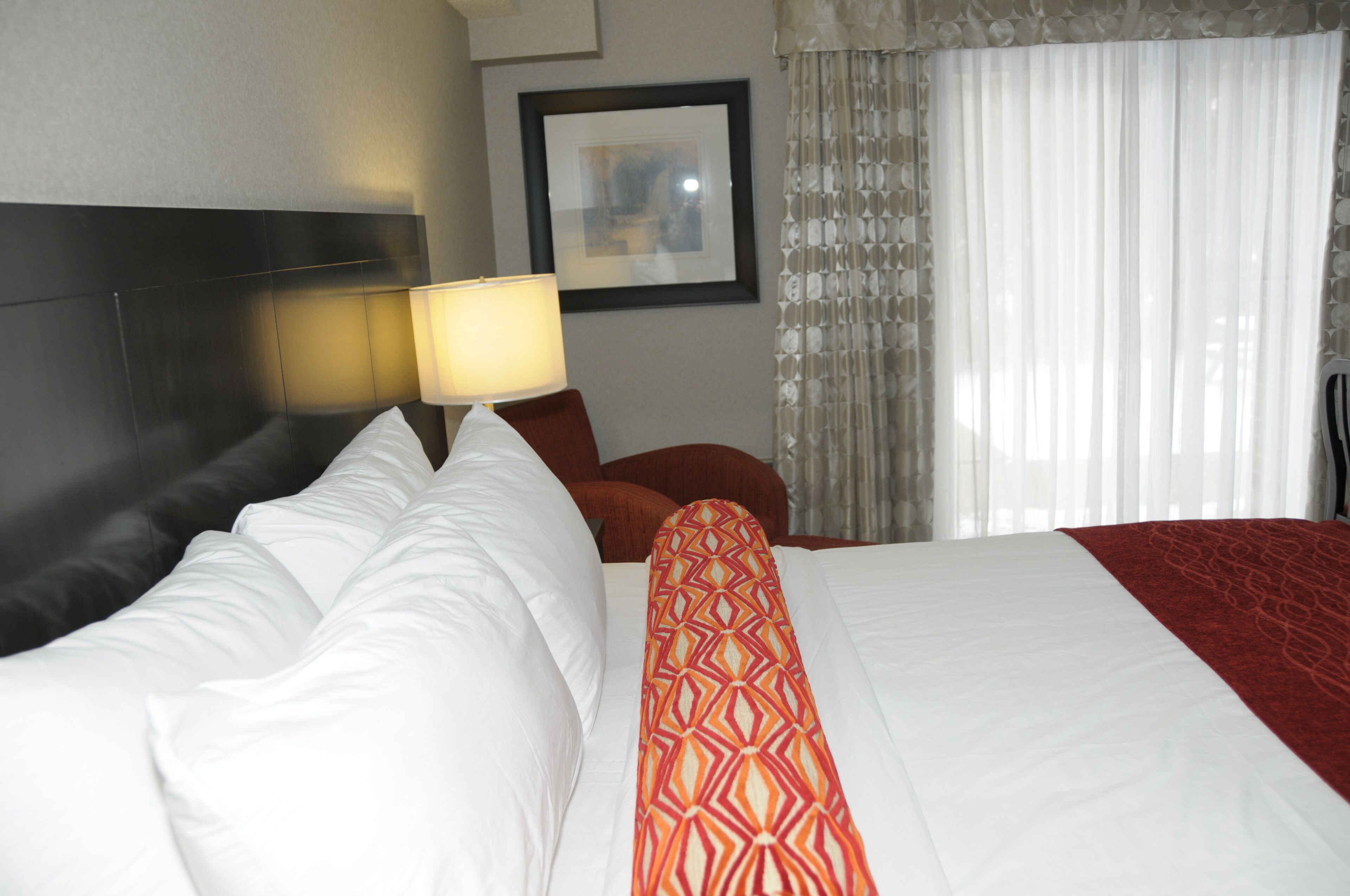 A hotel room with a neatly made bed, a headboard, a lamp, a framed picture on the wall, and a window with curtains.
